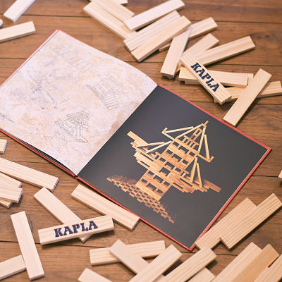 Open red Kapla art book showing building designs, surrounded by light wooden planks on a wooden floor.