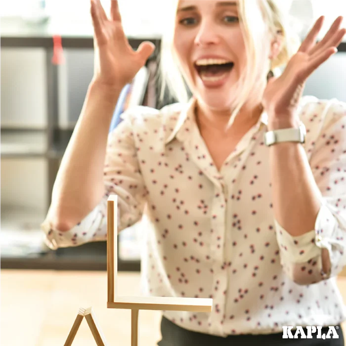 Excited woman cheers behind a precarious structure of thin wooden planks. Kapla logo in the corner.