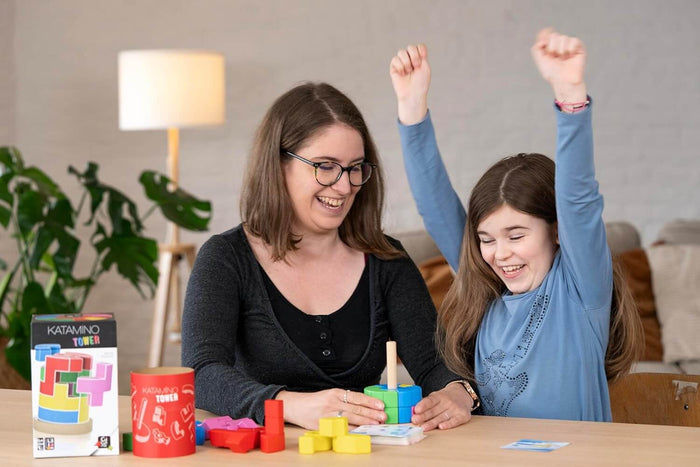Woman and girl celebrate building a colorful wooden block tower; the game box and red canister sit on the table.