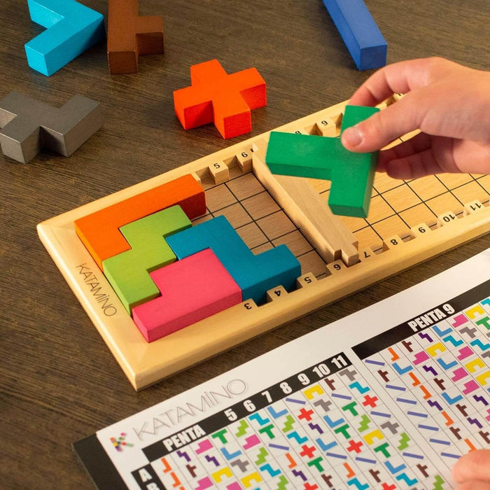 Hand placing a green wooden block into a Katamino board with colorful geometric shapes and a puzzle guide below.