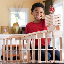 Smiling boy drops a red ball into an elaborate tower built from light-colored rectangular wooden planks.