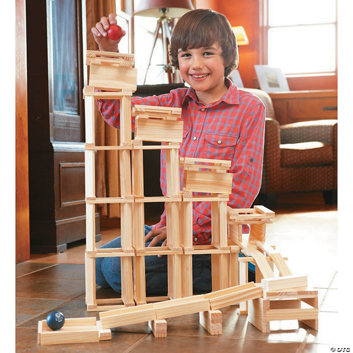 Smiling boy building a tall tower and ball run using light-colored rectangular wooden planks on a tile floor.