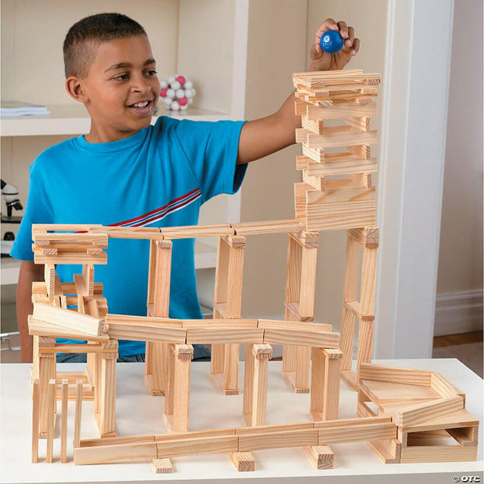 A boy in a blue shirt places a blue ball atop a tall tower and ramp system built from light-colored wooden planks.