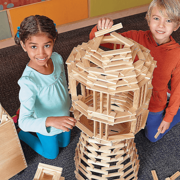 Two children building a tall, complex tower using hundreds of light-colored rectangular wood planks on a grey carpet.