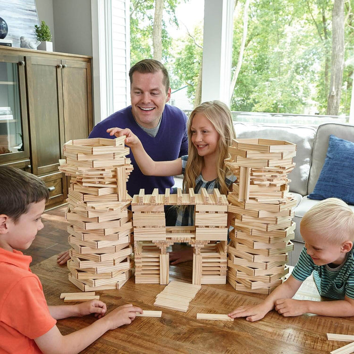 A family building a large castle structure with small rectangular natural wood planks on a wooden table.