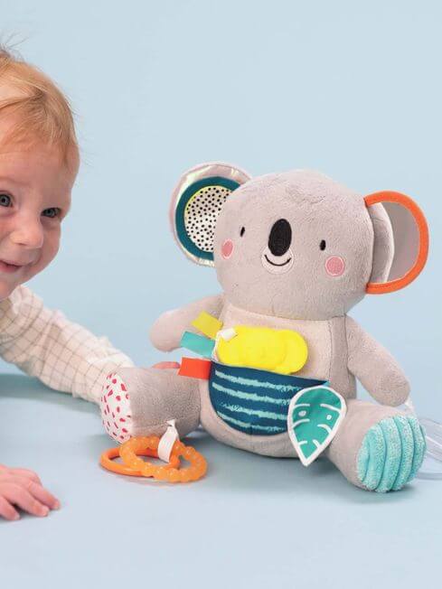 Grey plush koala with a mirror ear and colorful teethers, next to a smiling baby on a blue background.