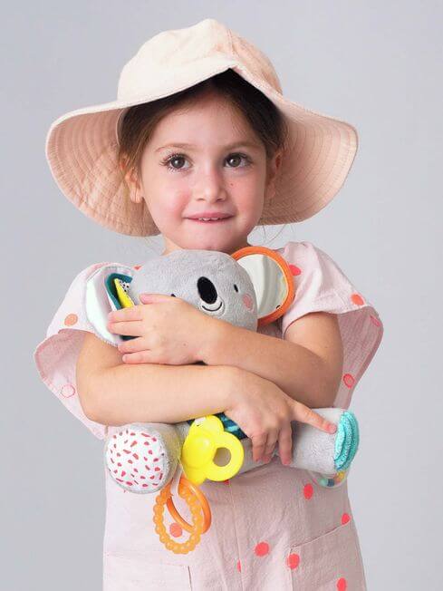 Girl hugs a gray plush koala toy with a mirror ear, patterned feet, and colorful dangling plastic rings and teether.