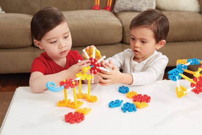 Two children building with chunky red, blue, and yellow plastic rods and rounded connectors on a white table.