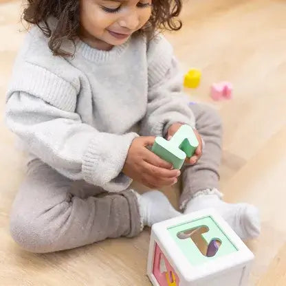 Toddler in a grey sweater holding a light green number block over a white shape sorter toy on a wood floor.