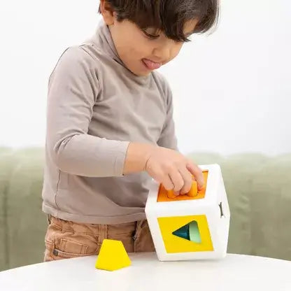 Toddler inserting colorful geometric blocks into a white shape sorter cube with yellow and orange panels.