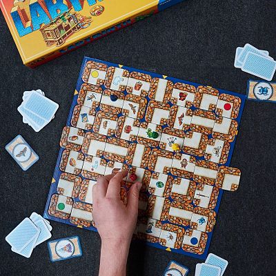 Hand moves a tile on a Labyrinth board game grid. Features colorful pawns, blue cards, and a yellow box.