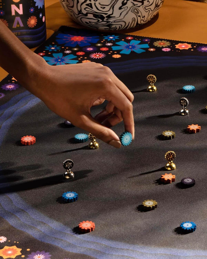 Hand picks up a blue gear token from a dark mat with colorful wooden gears and gold and silver metallic flower pawns.
