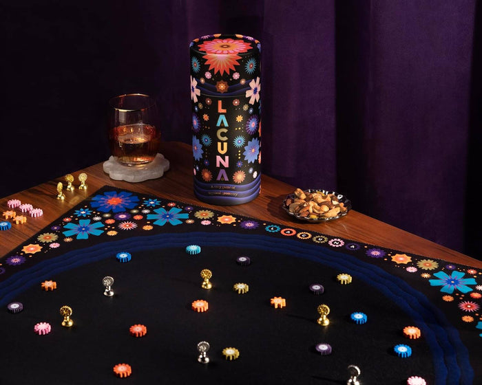 Lacuna cylindrical floral tube and black playmat with colorful flower tokens and metallic pieces on a dark wooden table.