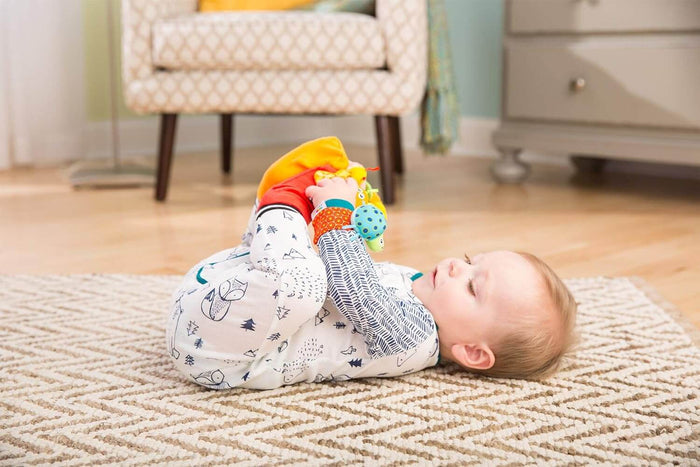 Baby on a rug playing with colorful plush bug-themed foot and wrist rattles in bright orange, yellow, and blue.