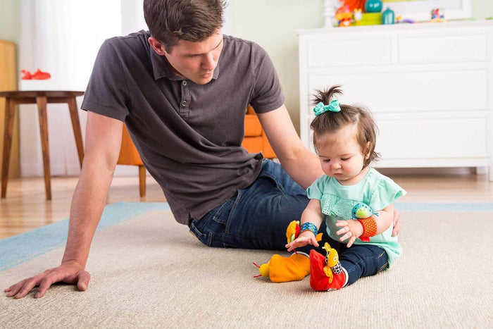 Baby girl wearing colorful plush bug-themed wrist and foot rattles while sitting on a rug with her father.