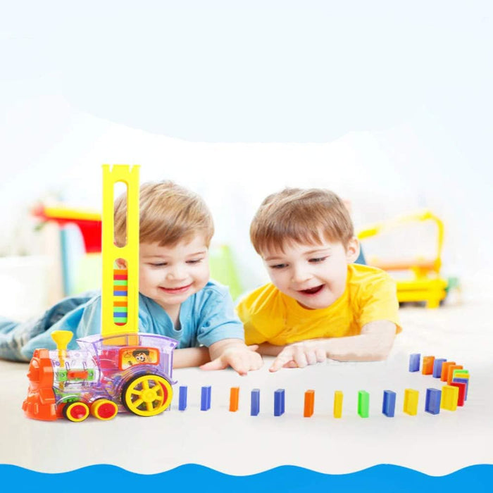 Two boys watch a colorful, transparent toy steam engine lay a line of upright, multi-colored dominoes.