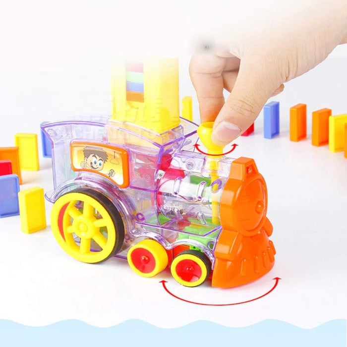 Hand turning the yellow chimney of a clear purple toy train with orange front, leaving a trail of colorful dominoes.