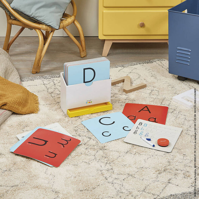 Colorful alphabet tracing cards, a wooden stand, and a writing board with marker on a rug in a playroom.
