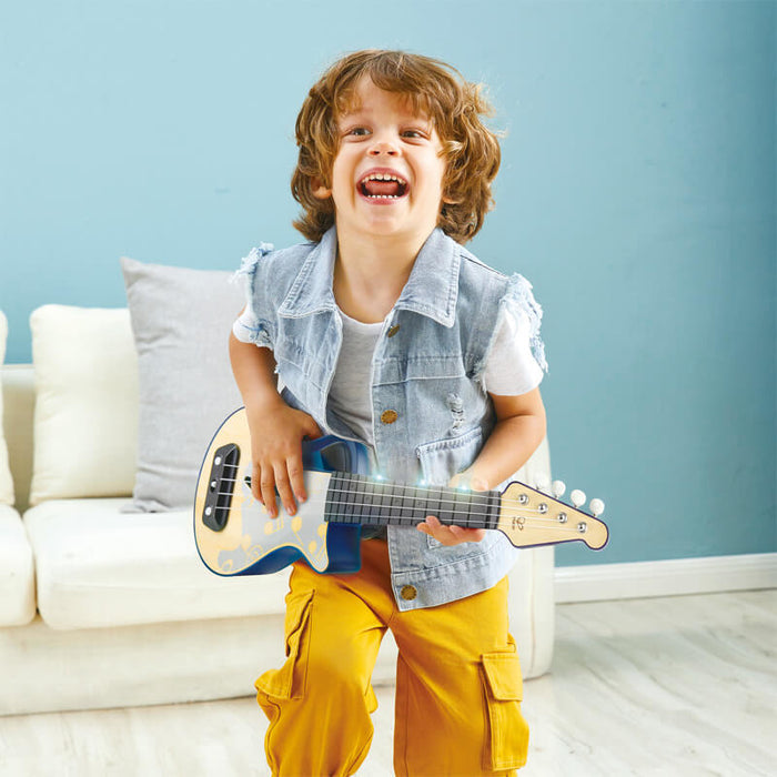 Young boy laughing while playing a blue and tan toy ukulele with light-up frets and music note graphics.