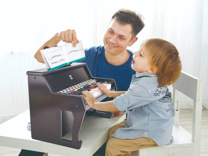 Toddler plays a black upright toy piano with light-up keys as a man holds an open music book.