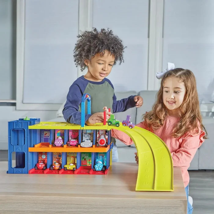 Children playing with a 3-level colorful toy garage, character cars, and a curved lime green ramp on a wooden table.