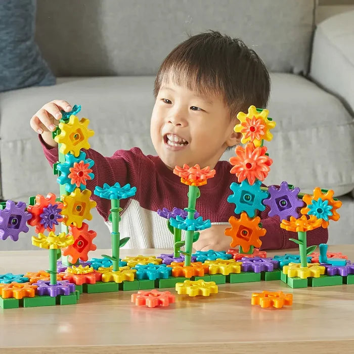 Smiling child building with vibrant orange, blue, and yellow flower gears and green stems on a wooden table.