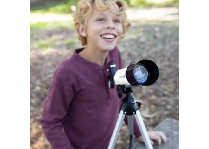 Smiling boy in a purple shirt next to a white and black telescope on a silver tripod in a sunny outdoor setting.