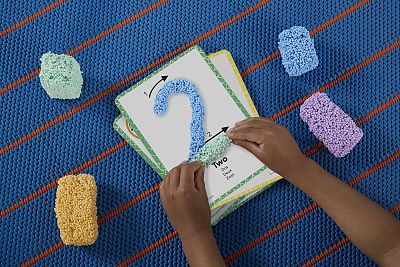 Child's hands molding colorful, textured foam into the number 2 on a learning card over a blue striped mat.