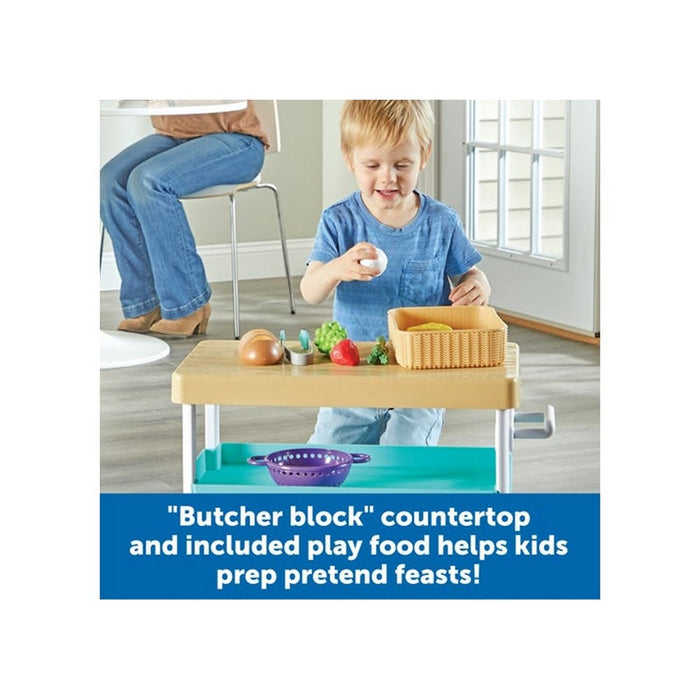 Child playing at a tan and teal toy kitchen island with play food, a basket, and a purple colander.