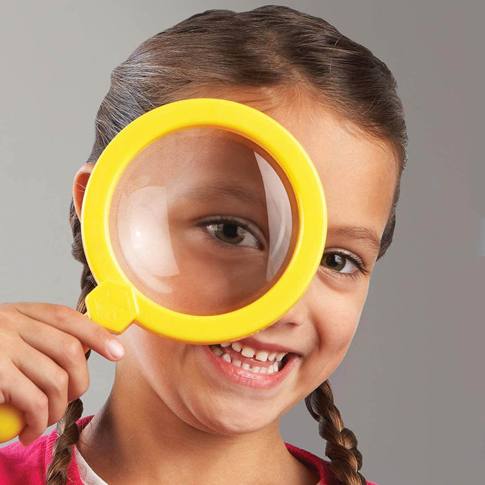 A smiling girl holding a jumbo yellow magnifying glass over her eye, making it appear larger.