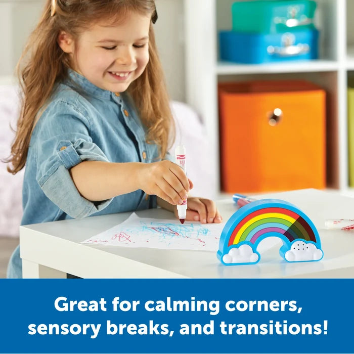 Smiling girl draws next to a blue rainbow-shaped visual timer with white clouds on a white table.