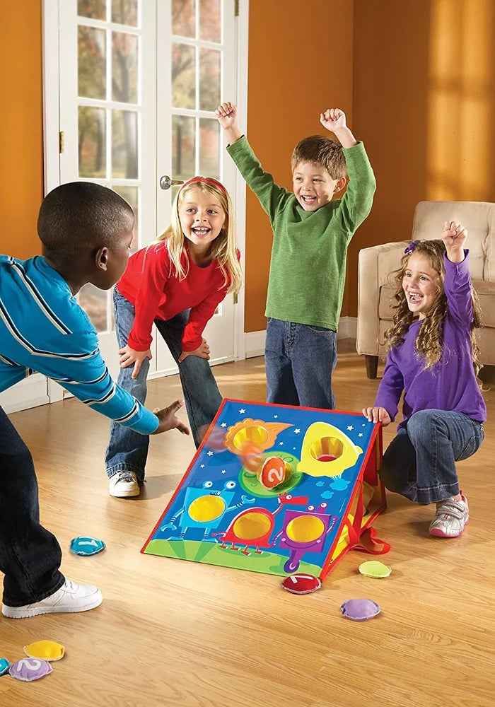 Four children play bean bag toss with a colorful slanted board featuring circular holes and scattered bean bags.