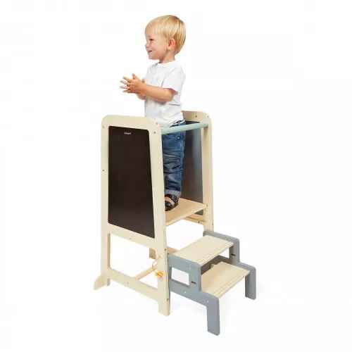 Toddler standing on a wooden learning tower with grey steps, a safety rail, and an integrated chalkboard.