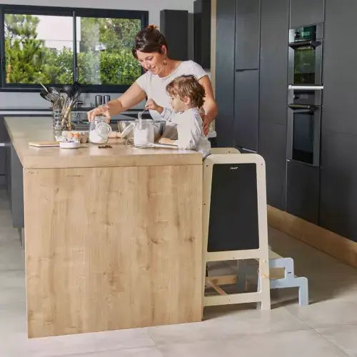Light wood toddler tower with a black chalkboard back, shown with a child and adult at a kitchen counter.