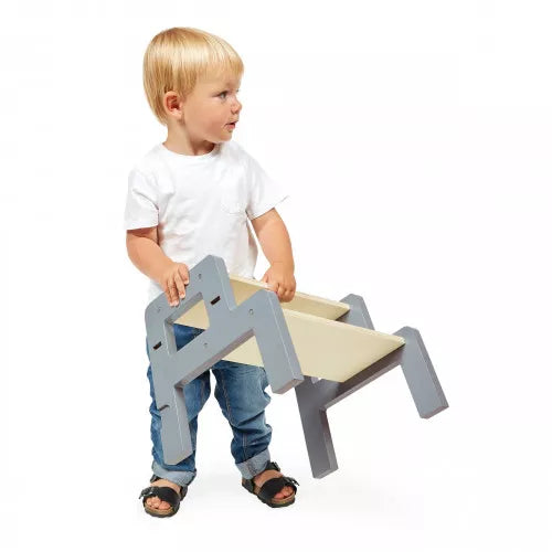 A toddler in a white shirt and jeans lifts a gray and light wood step stool on a plain white background.