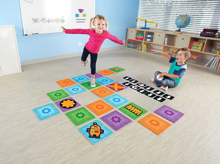 Children play on colorful square floor mats with gear and arrow symbols to build a coding path in a classroom.