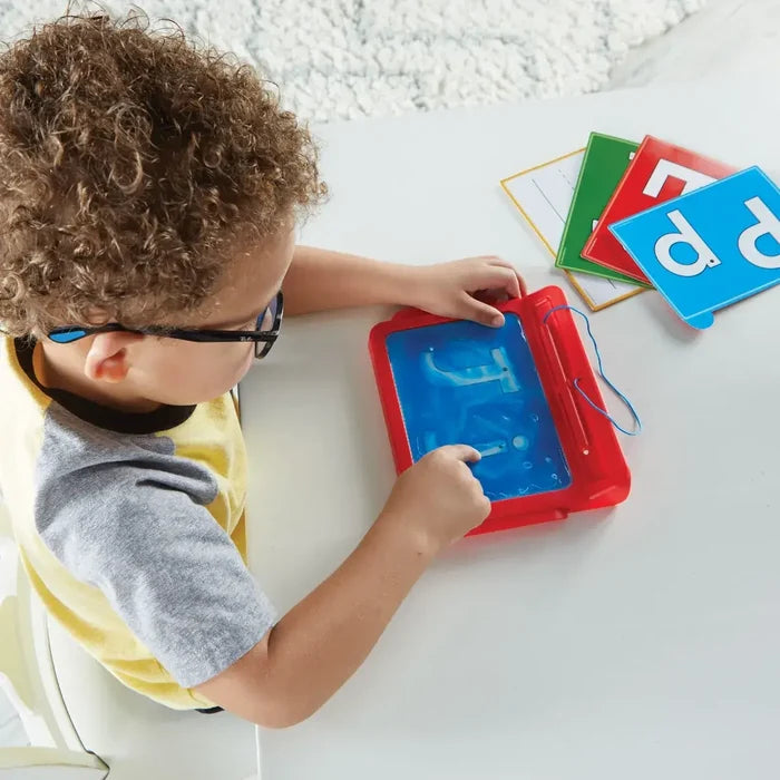 Child traces the letter J on a red gel-filled pad next to colorful alphabet cards on a white table.