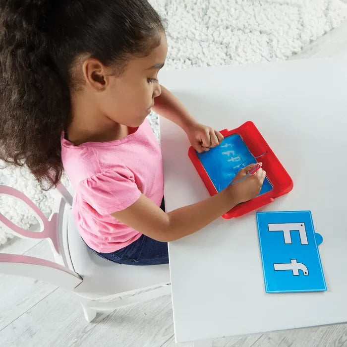 Child uses a stylus to trace letters on a blue sensory gel pad in a red tray next to a letter card.