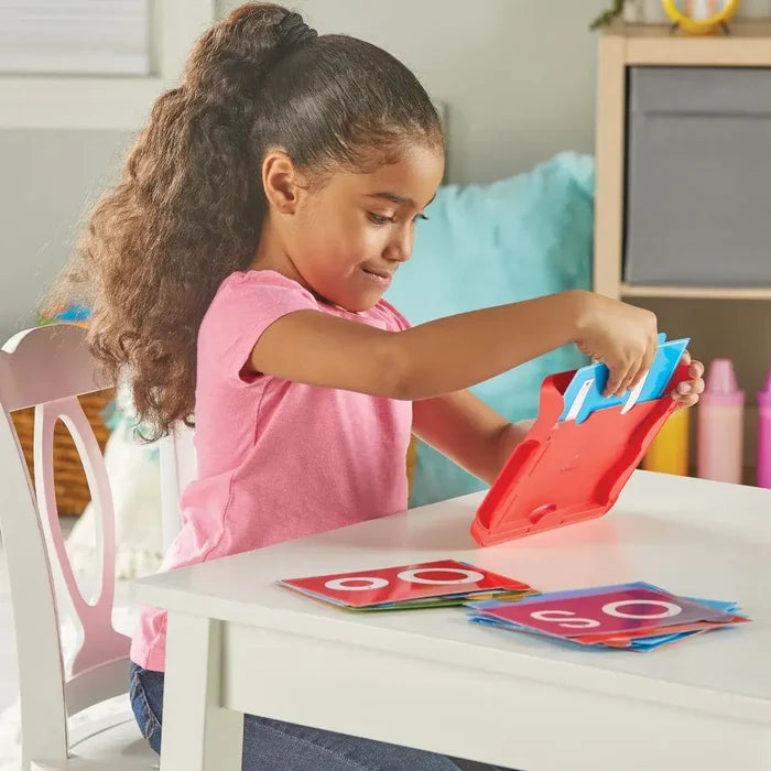 A girl slides a blue letter card into a red sensory pad frame next to stacks of colorful flashcards.