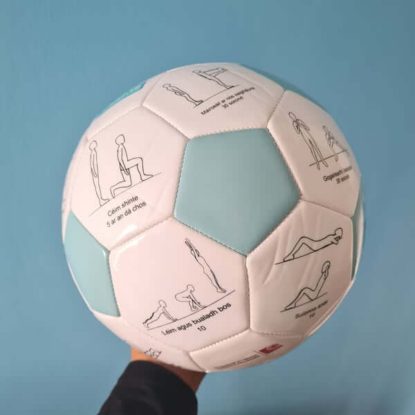White and light blue soccer ball with exercise diagrams and Irish text, held by a hand against a blue background.
