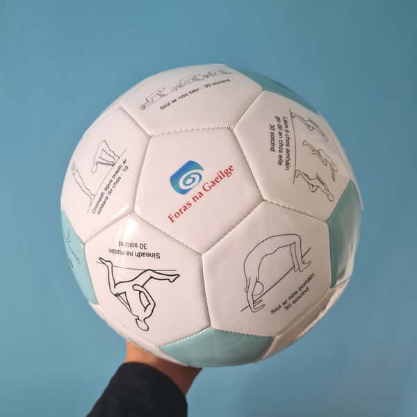 White and teal soccer ball with illustrations of stretches and Irish text, held against a blue background.
