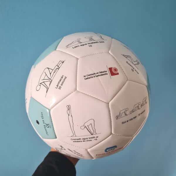 White soccer ball with light blue panels featuring black line drawings of exercise poses and Irish text.