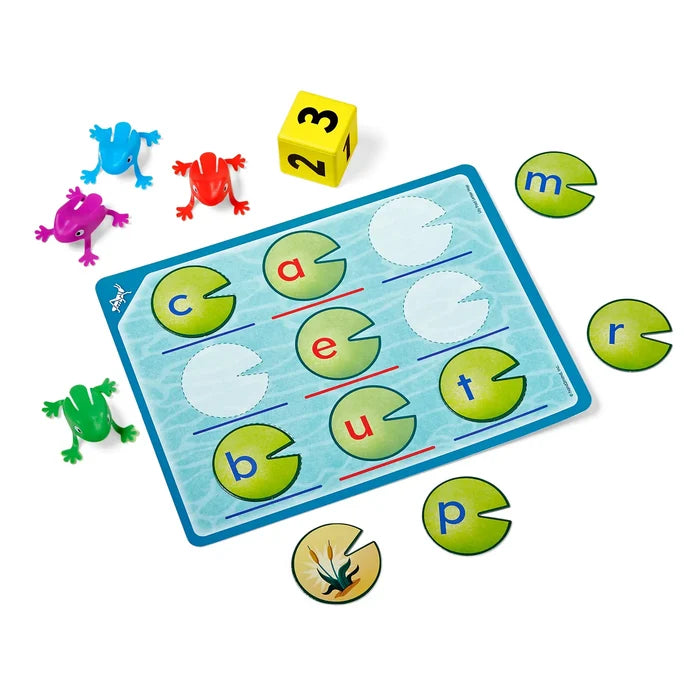 An educational literacy game featuring a game board, letter lily pads, colorful frog figurines, and a yellow numbered die.
