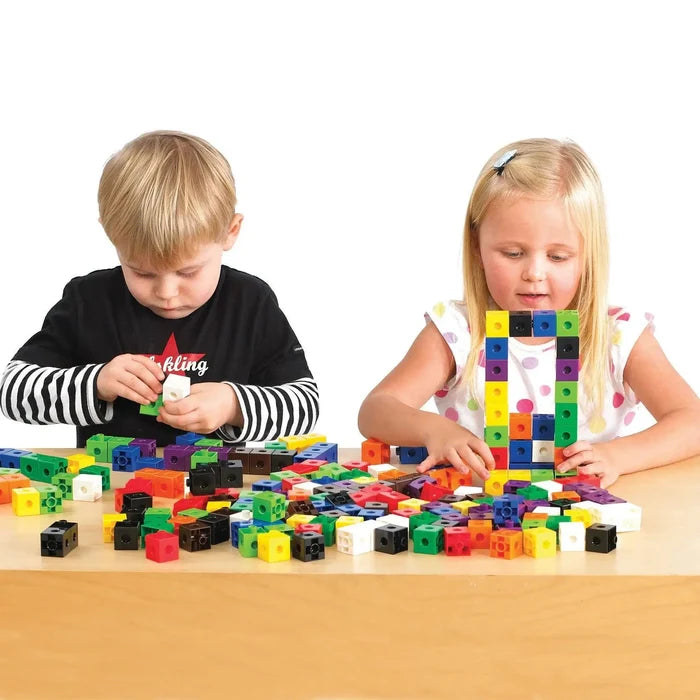 Two children building structures with multi-colored interlocking plastic cubes on a light wood table.