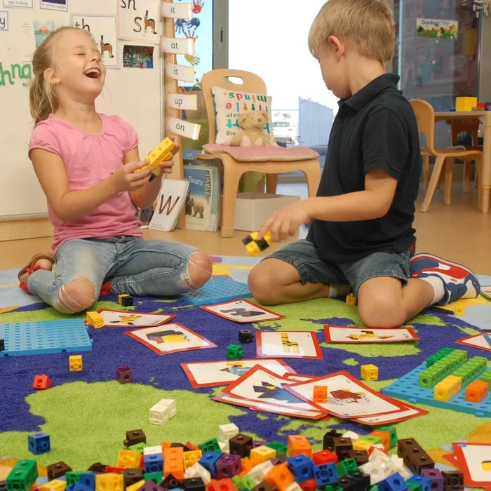 Children using multicolored linking cubes, blue baseplates, and activity cards on a world map rug in a classroom.