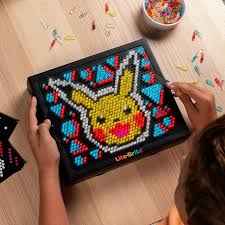 Hands holding a Lite-Brite board with a glowing Pikachu mosaic made of yellow, red, and blue pegs on a wood table.