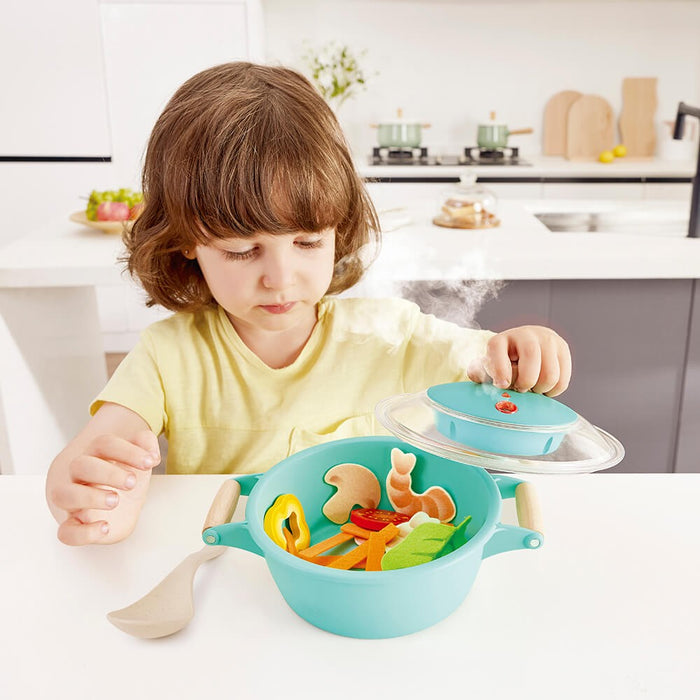 Young girl lifts the lid of a teal toy pot with play food as realistic mist rises from the center.