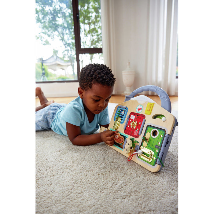 Young boy on a rug plays with a house-shaped wooden activity board featuring colorful doors and metal latches.