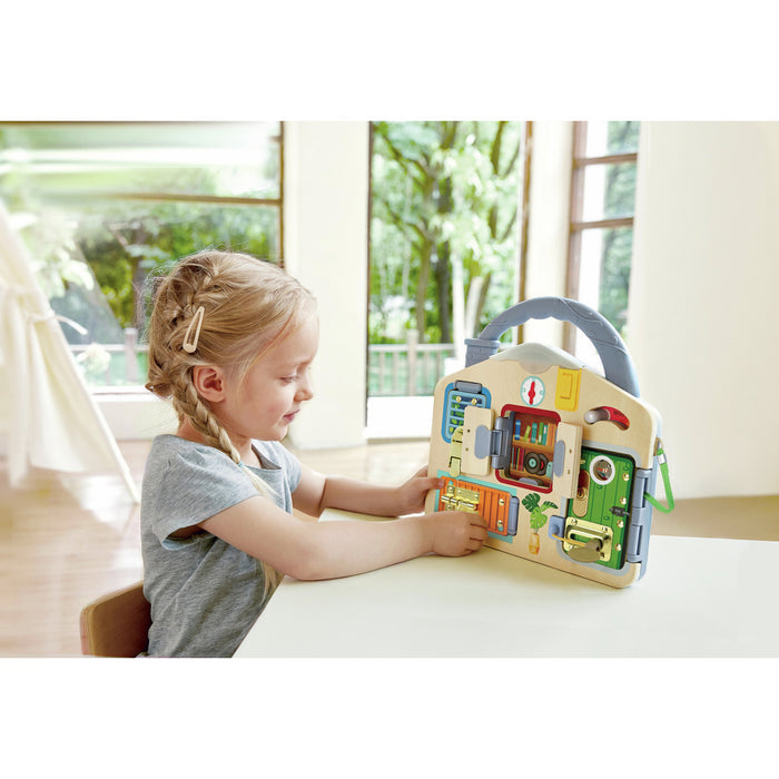 Blonde girl at a table plays with a wooden house activity board featuring colorful doors and metal latches.