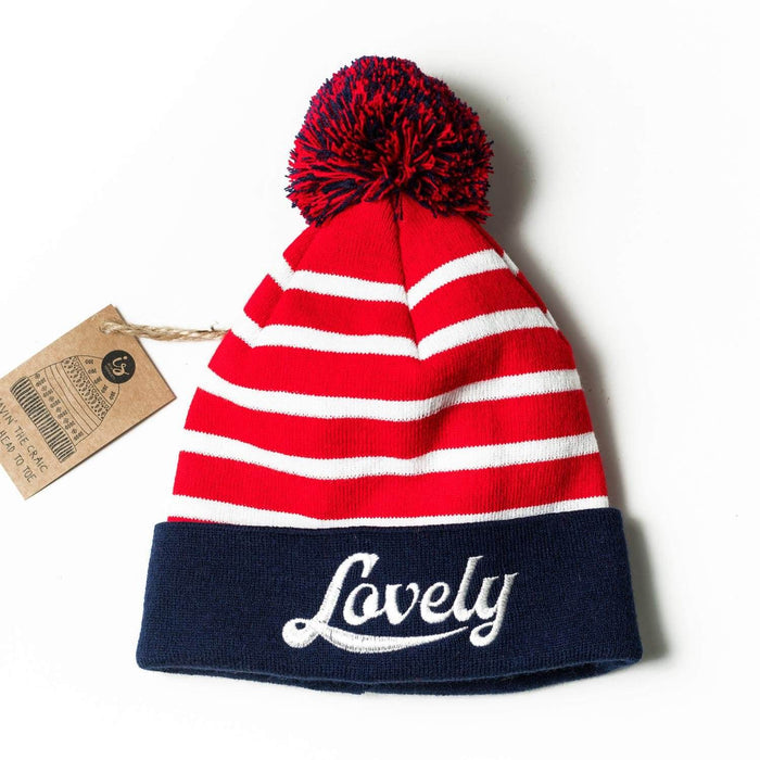 Adult knit hat with red and white stripes, a blue brim embroidered with Lovely, and a navy and red pom-pom.